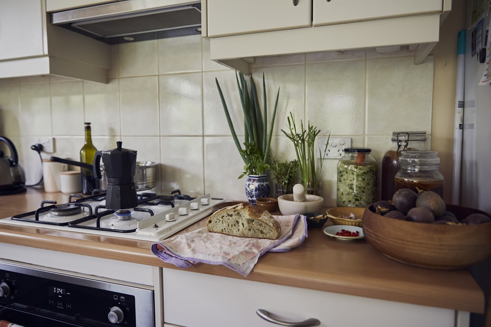 A permaculture kitchen bench scene, showing sourdough, pickles, homegrown figs and veggies, and coffee bubbling on the stovetop. It's the home office of someone who applies permaculture in business.