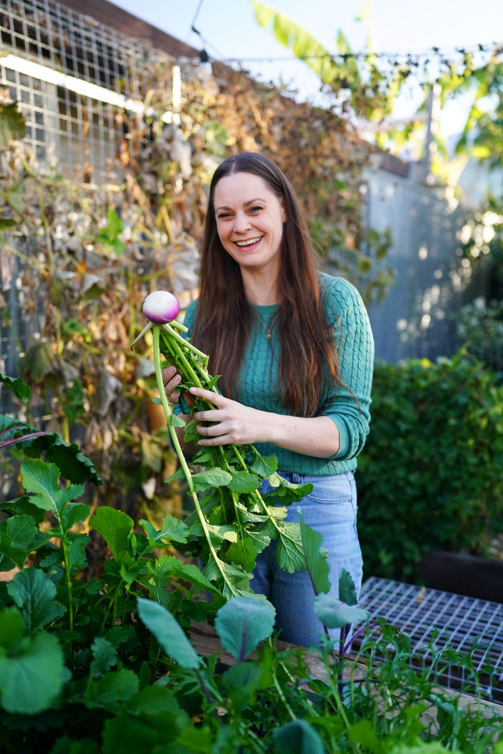 Koren Helbig standing in her small city permaculture garden, smiling at the camera and holding a freshly picked homegrown turnip.