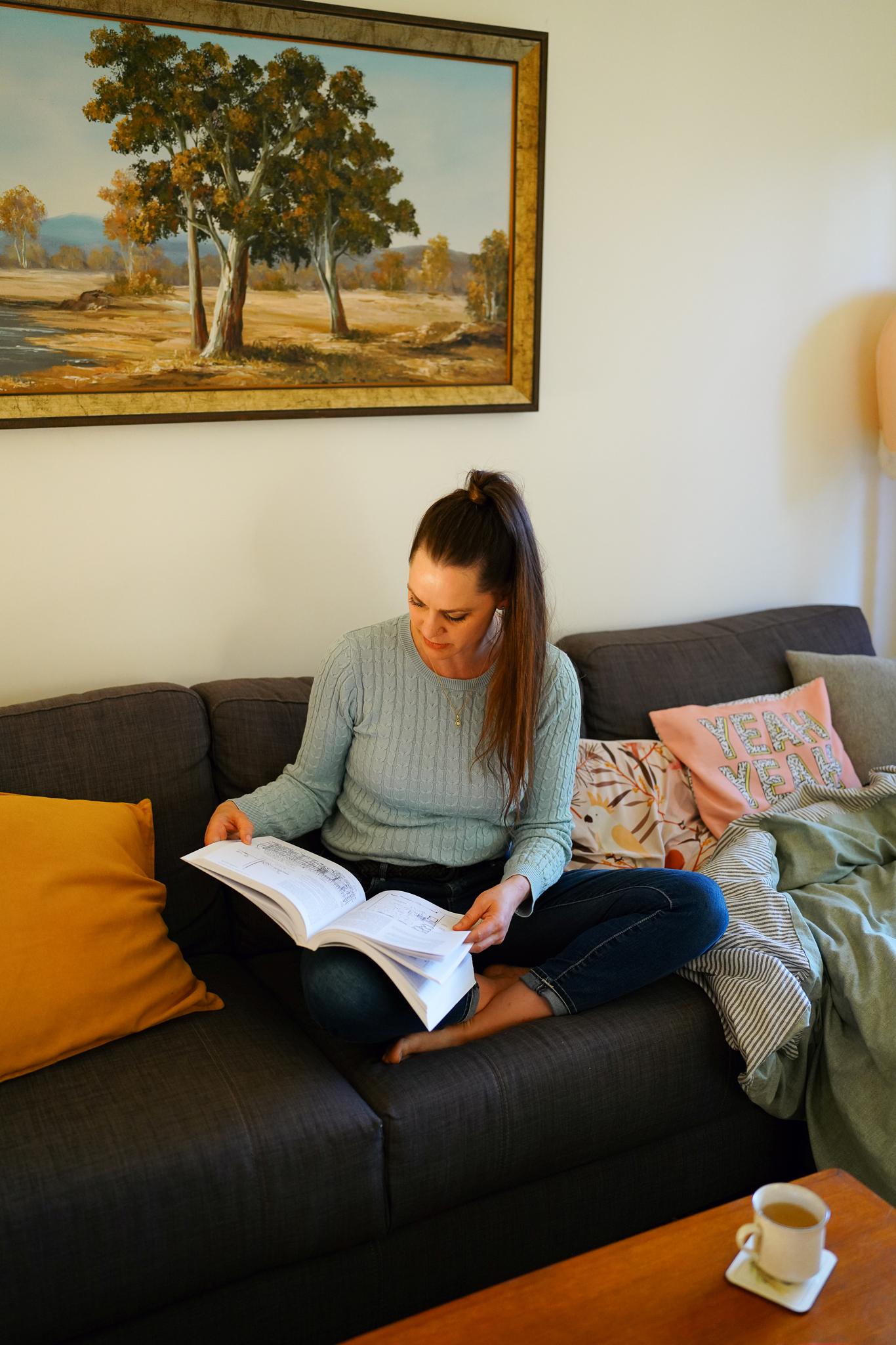 Koren sitting on her couch, reading a book, surrounded by comfy cushions and a blanket.