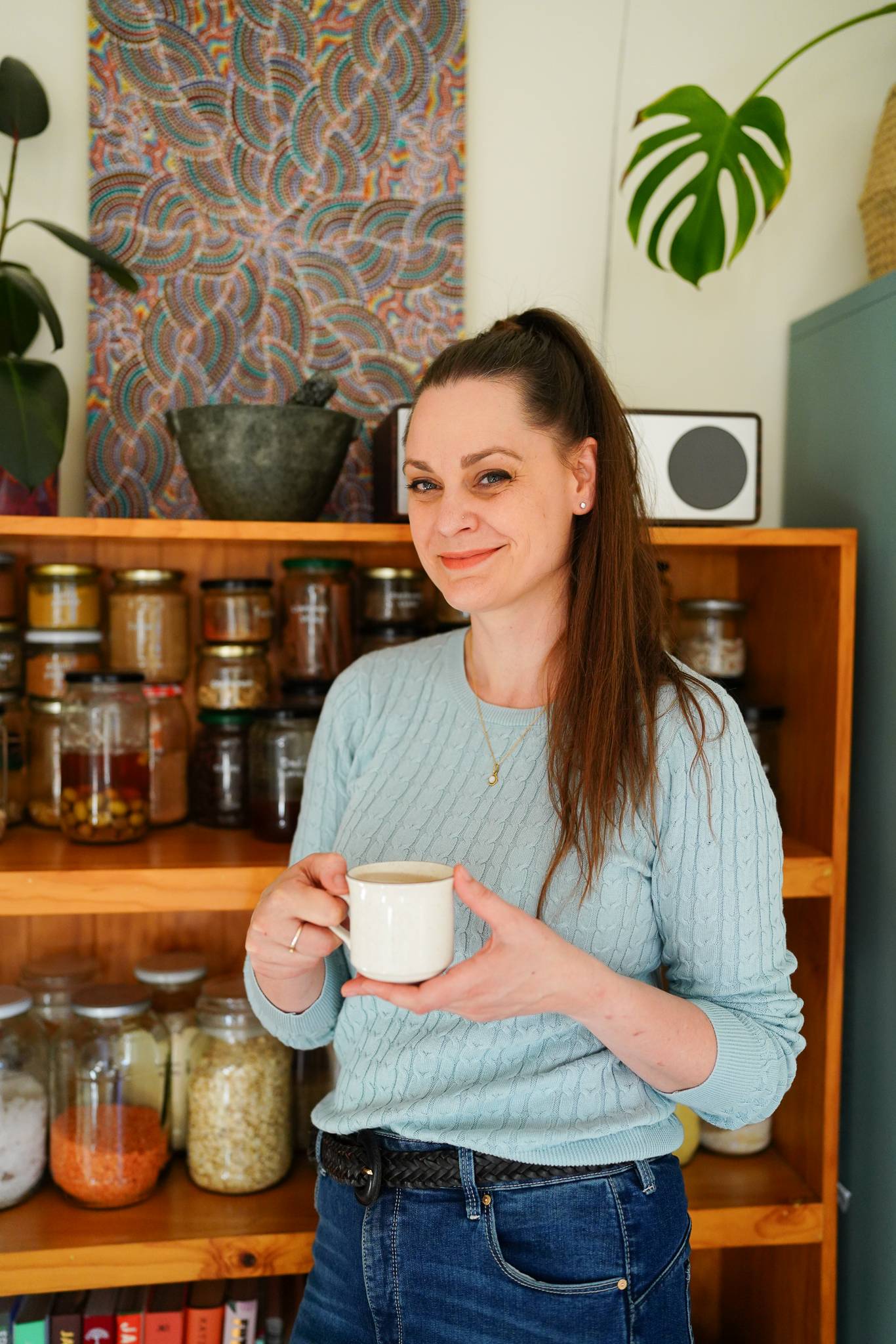 Koren Helbig standing in her kitchen, holding a cup of herbal tea. Behind her is a bookshelf full of jars of food.
