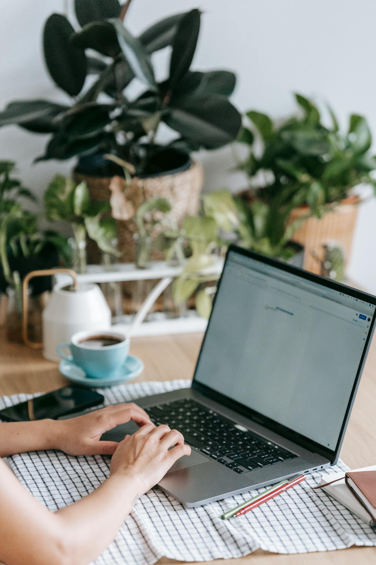 A laptop sitting atop a wooden table, surrounded by dark green pot plants, with a woman's hands typing on the keyboard.